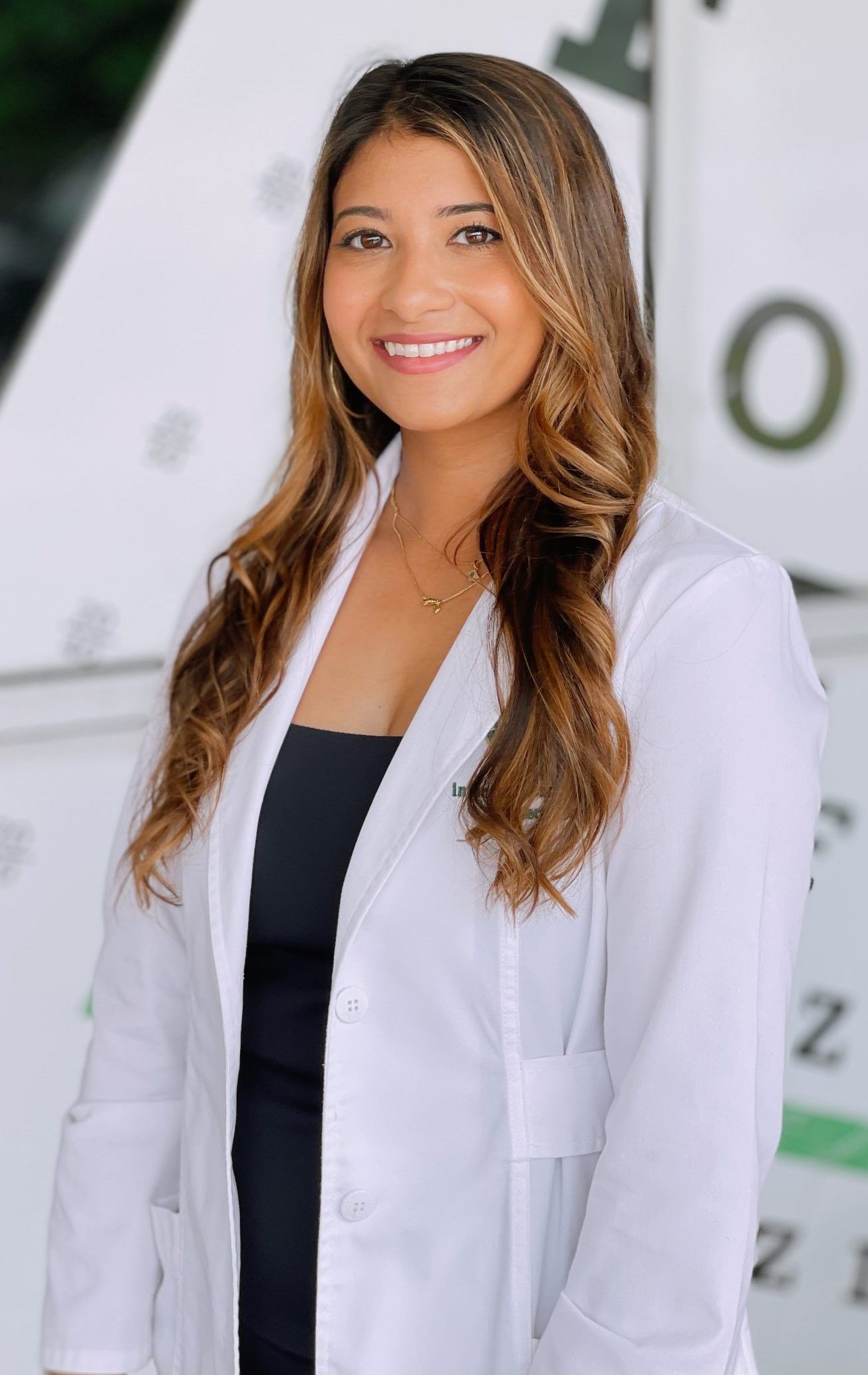 A woman in a white coat is smiling in front of an eye chart.