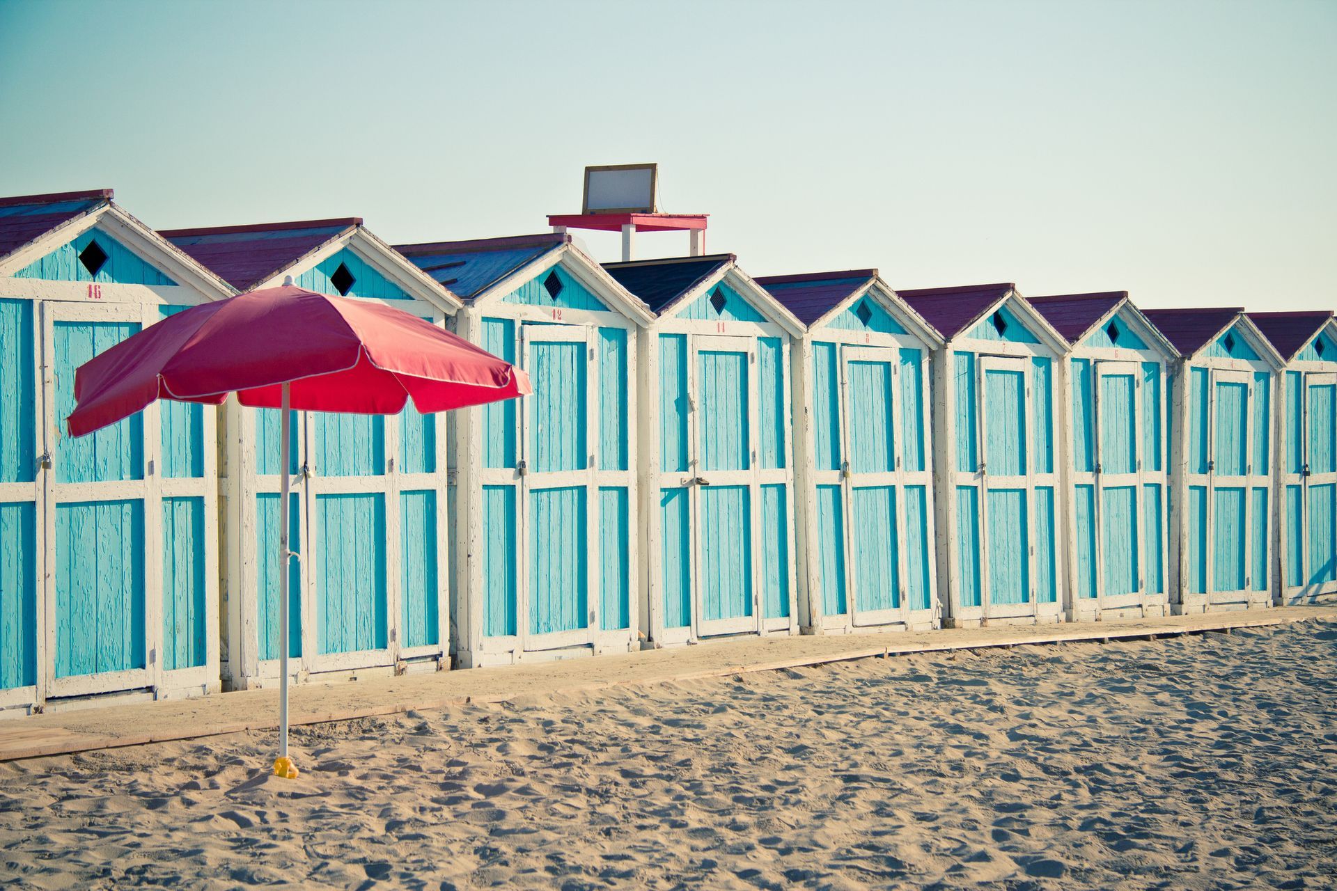 A red umbrella stands in front of a row of blue beach huts.