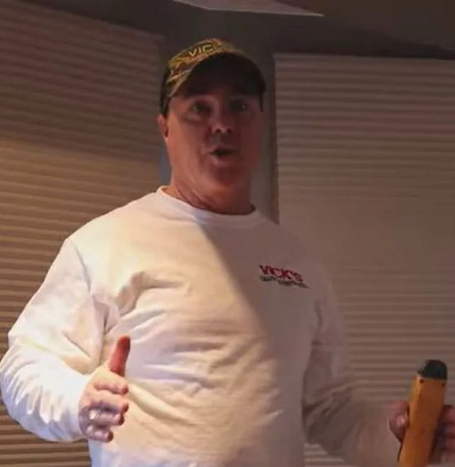Man in white shirt and camo hat speaking, holding a tool, standing indoors.