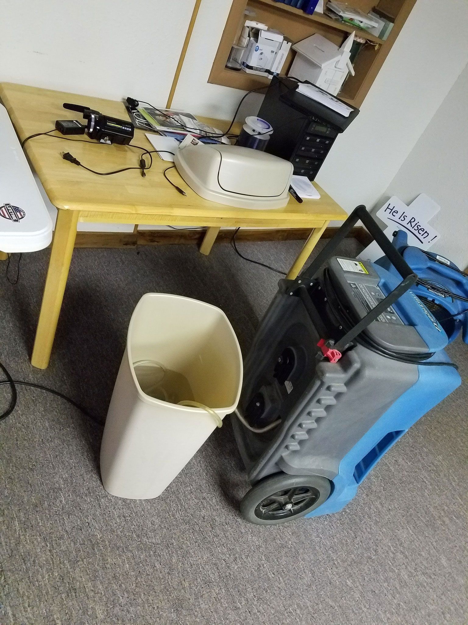 Beige trash can and dehumidifier next to a wooden table with a printer and office supplies on carpet.