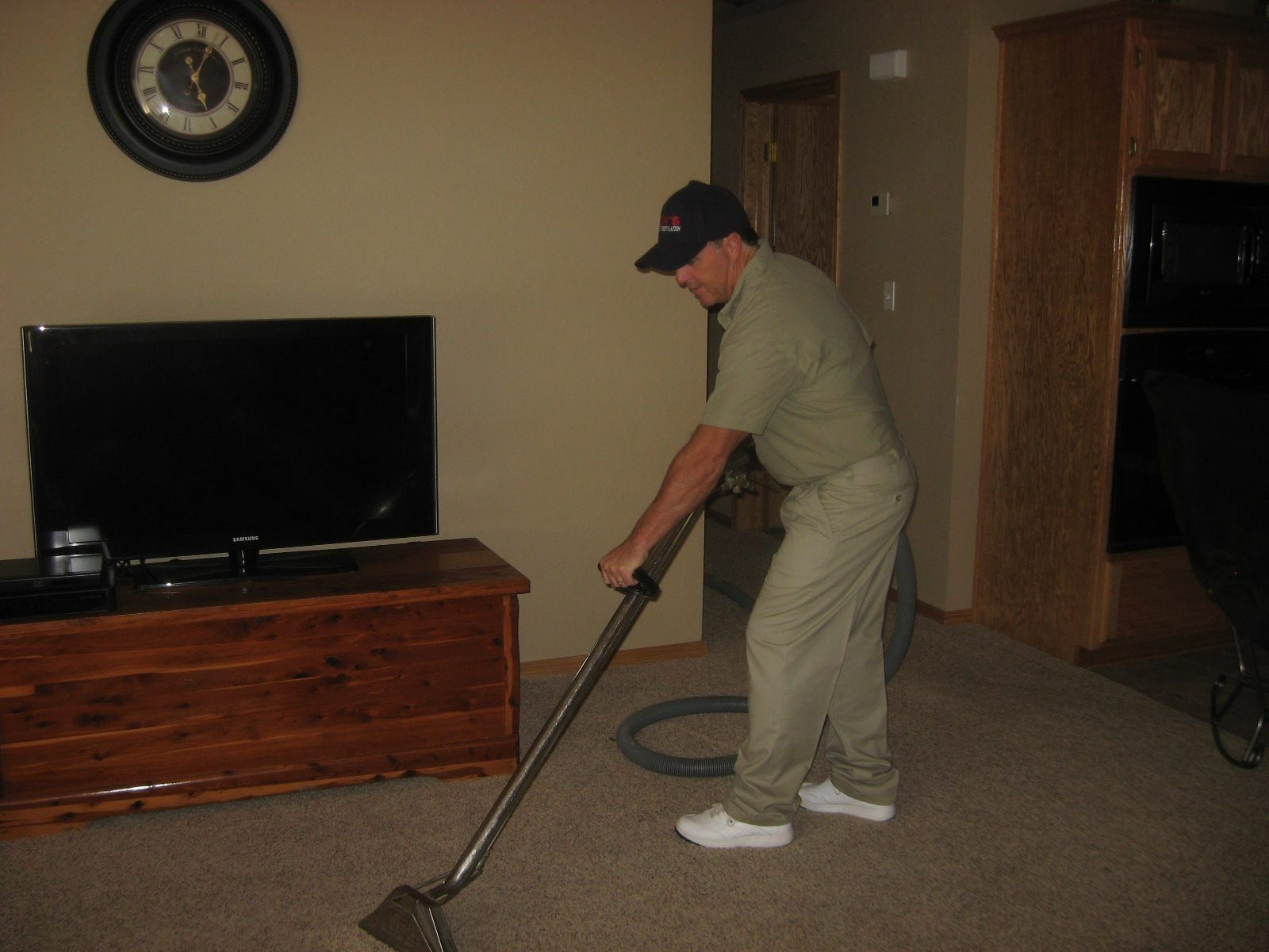 A man in a cap vacuums carpet in a living room, near a TV and wooden chest.
