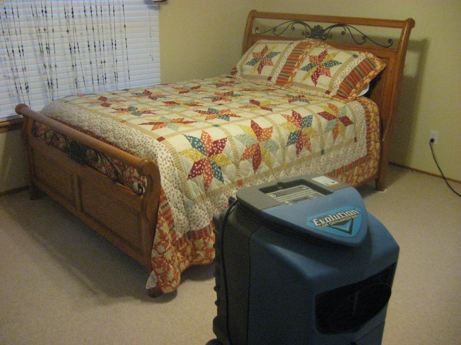 A bedroom with a wooden bed and a blue air purifier on the carpet. The bed has a floral quilt and pillows.