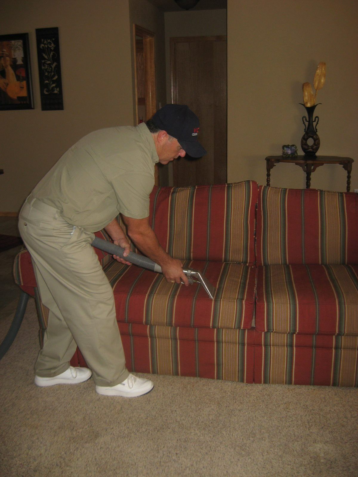 Man in khaki cleaning a red striped sofa with a vacuum in a living room.