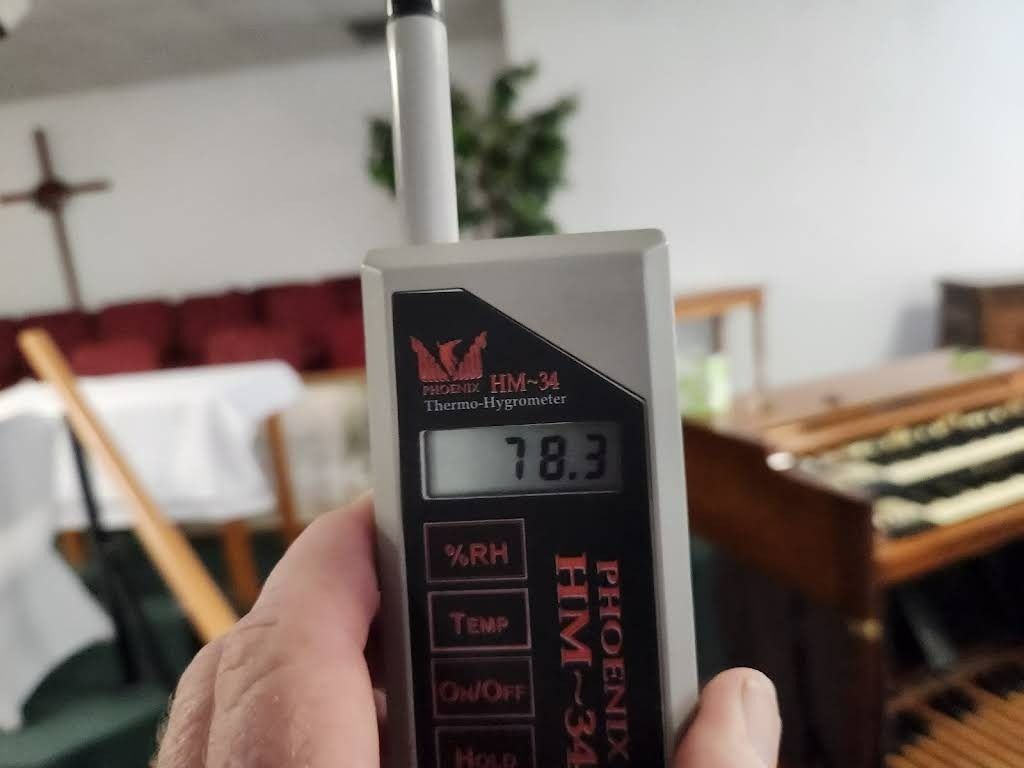 Hand holding a Phoenix HM-34 humidity meter displaying 78.3% in a church near an organ and altar.