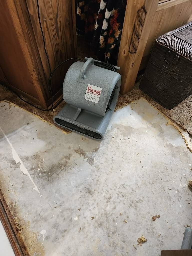 Gray air mover fan on a stained concrete floor, positioned near a wooden cabinet and a woven basket.