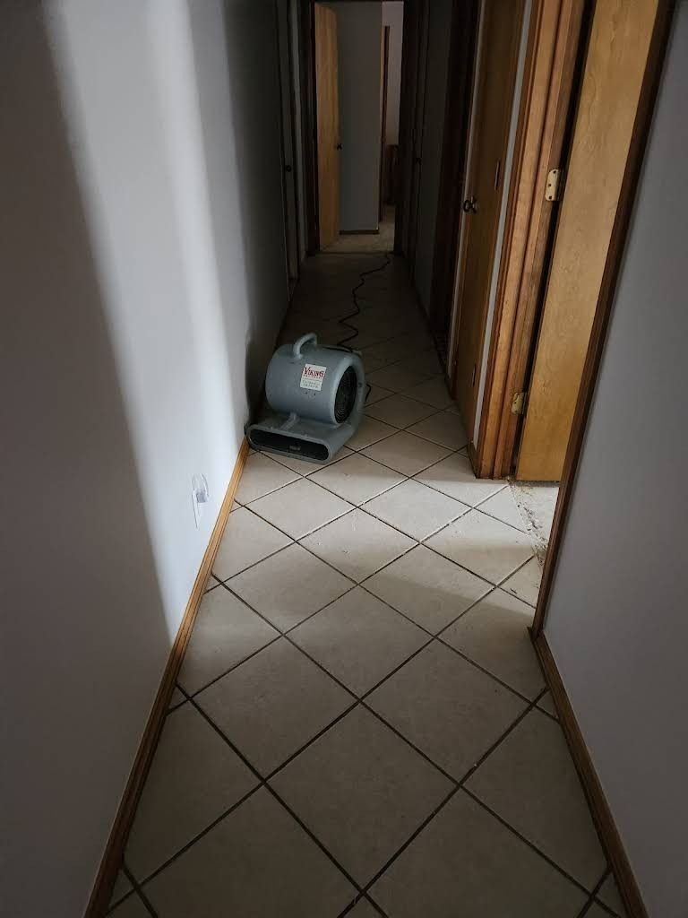 Fan in a hallway blowing air, drying the floor. Light tiles, doors on the right, hallway leads to a dark room.