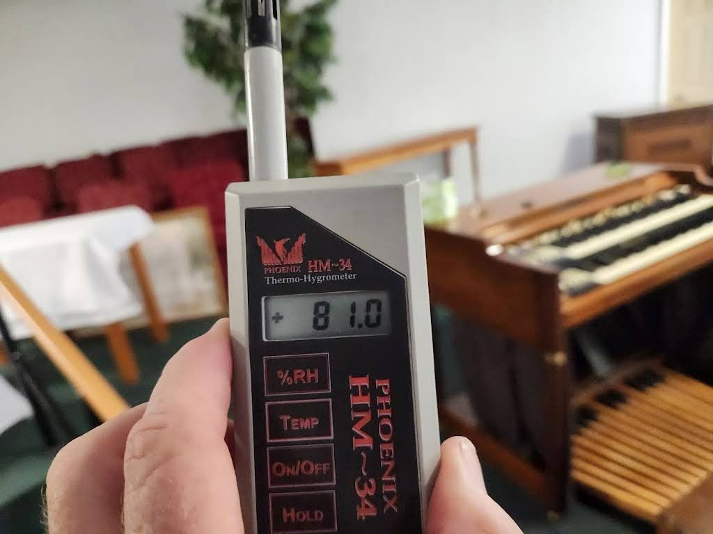 Hand holding a Phoenix HM-34 meter, displaying 81.0. An organ and furniture are in the background.