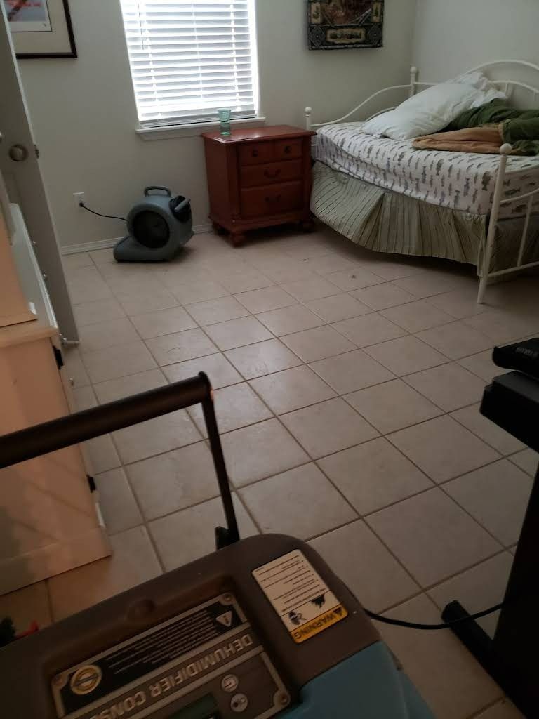 A room with a dehumidifier and fan; a bed, window, and nightstand are visible.