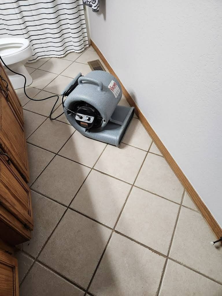 A gray industrial fan drying a corner of a bathroom with tile floor and white walls.