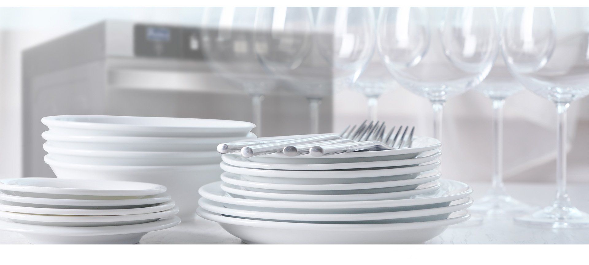 A stack of white plates , bowls , and wine glasses on a table.