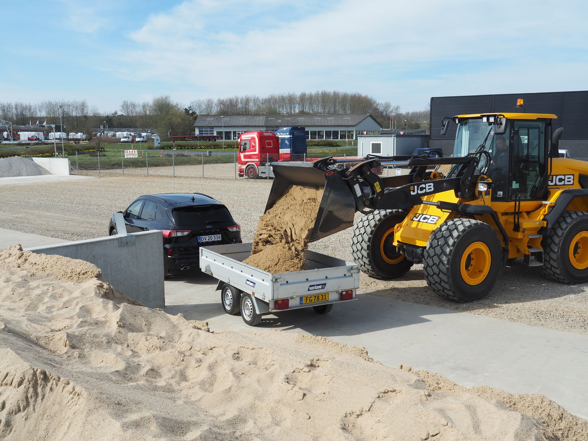 Gul JCB-læsser tømmer sand i en trailer trukket til en sort SUV udendørs.