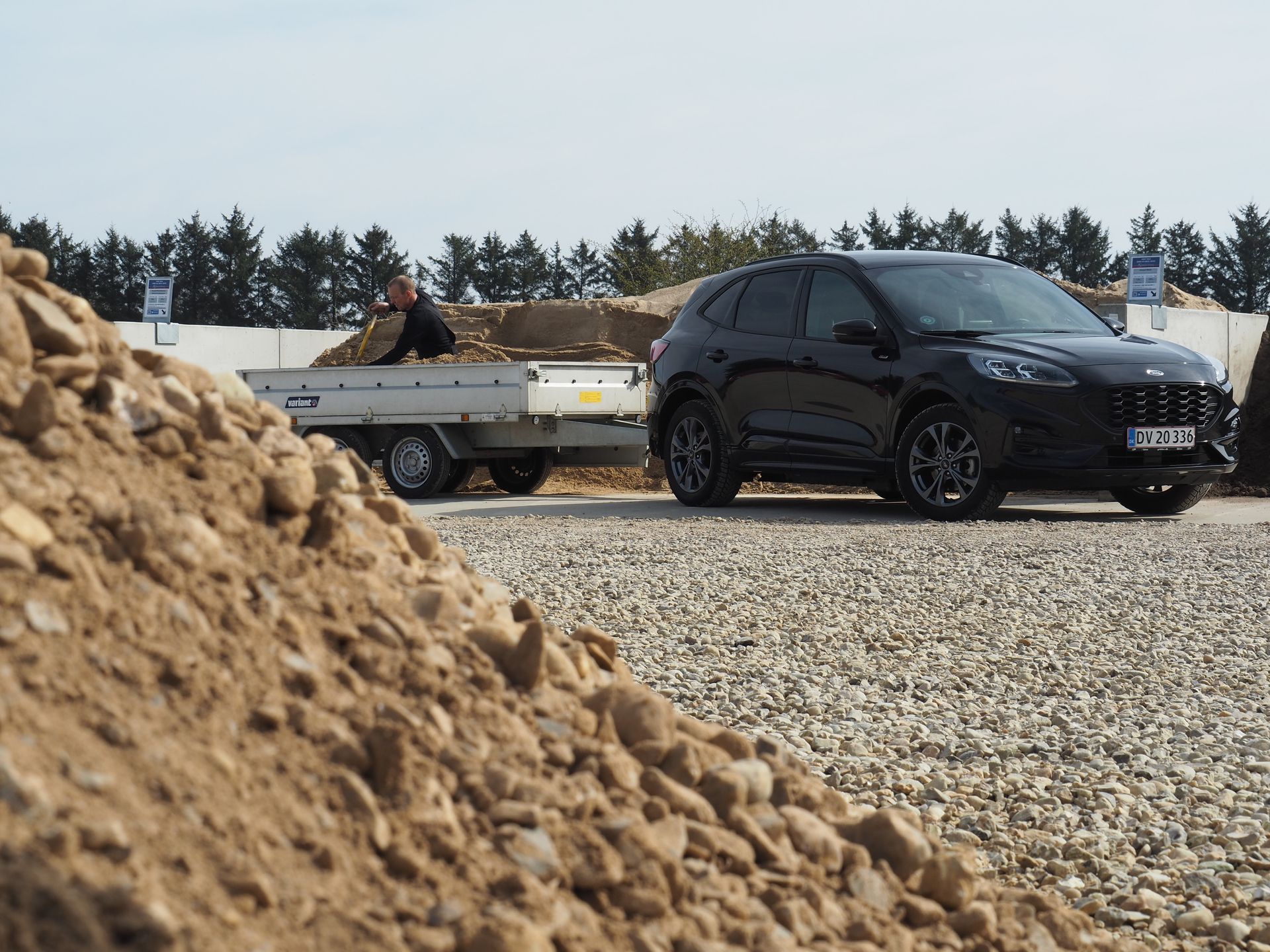 Sort SUV trækker en trailer læsset med grus på en grusplads, person i baggrunden.