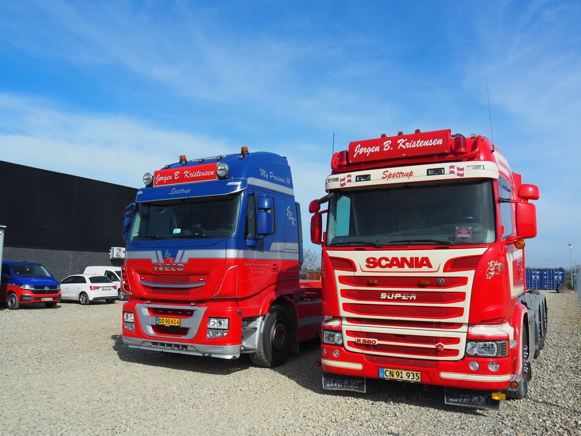 To røde og blå semitrailere parkeret udendørs på grus med en delvist overskyet himmel.