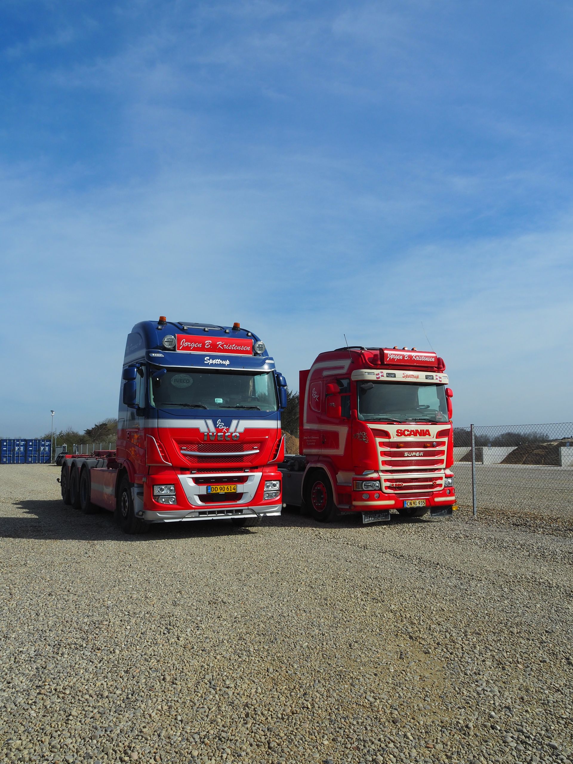 To farverige semitrucks parkeret på grus under en blå himmel; en rød og en blå.