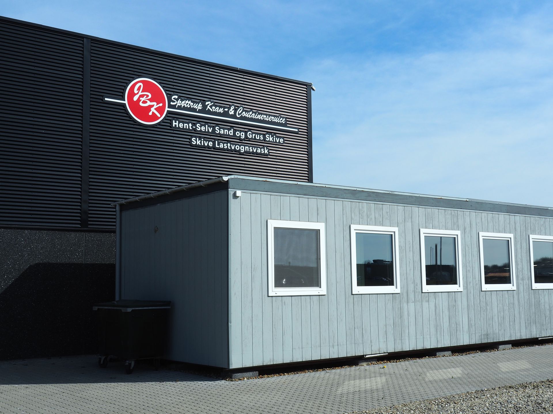 Gr&aring;t skur foran sort bygning med virksomhedens logo og tekst om sand, grus og lastvognsvask.