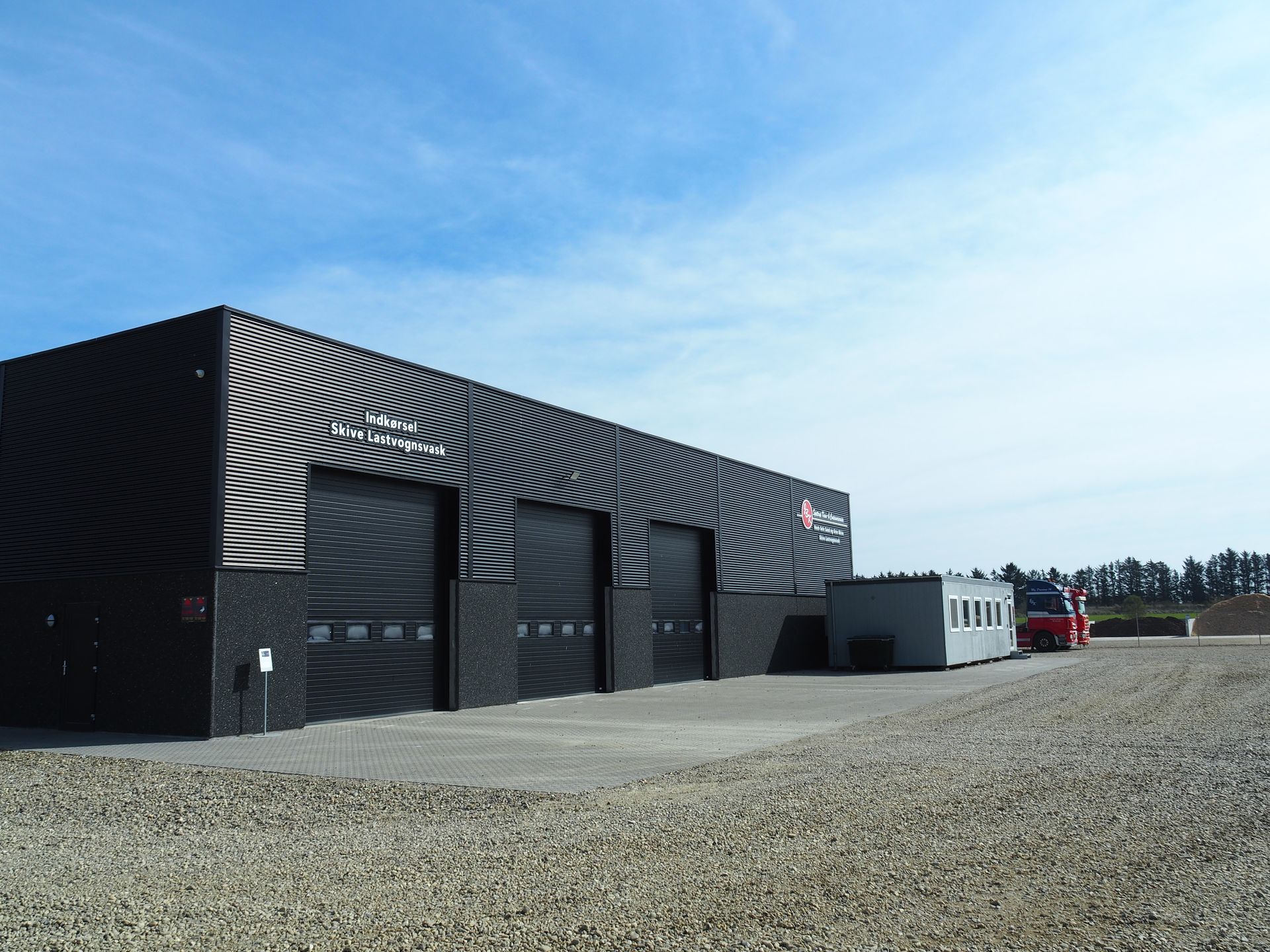 Sort industribygning med tre garageporte; grusgrund, blå himmel.