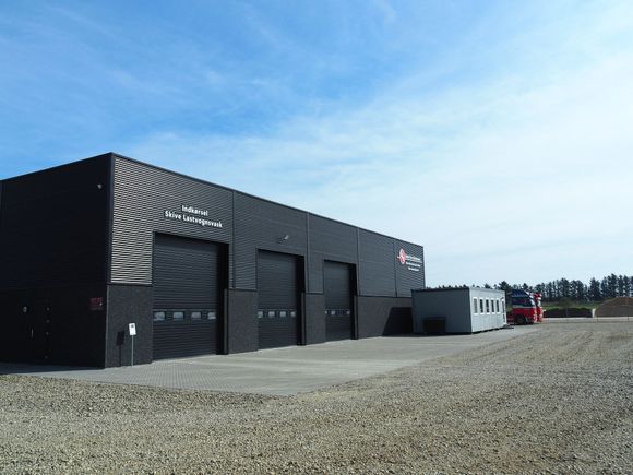 Sort industribygning med tre garageporte; grusgrund, blå himmel.