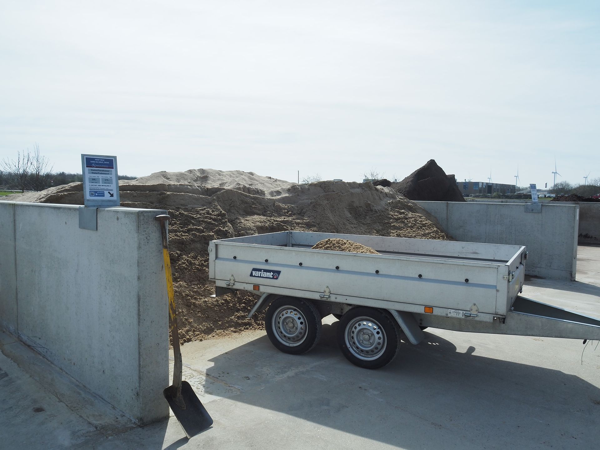 En trailer parkeret ved siden af ​​en betonvæg, læsset med kartofler, nær en bunke jord og skiltning.