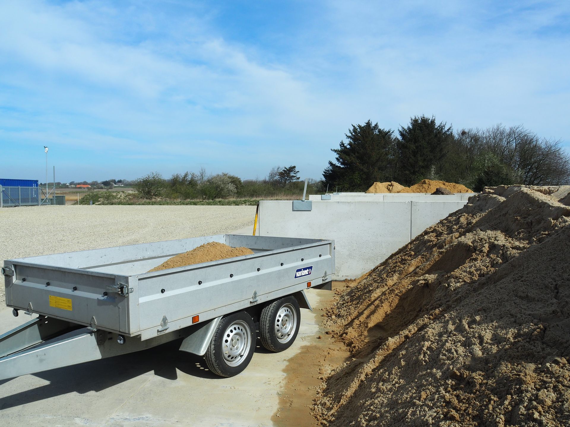 En sølvfarvet trailer læsset med sand, ved siden af ​​en betonvæg og sandbunker, under en blå himmel.