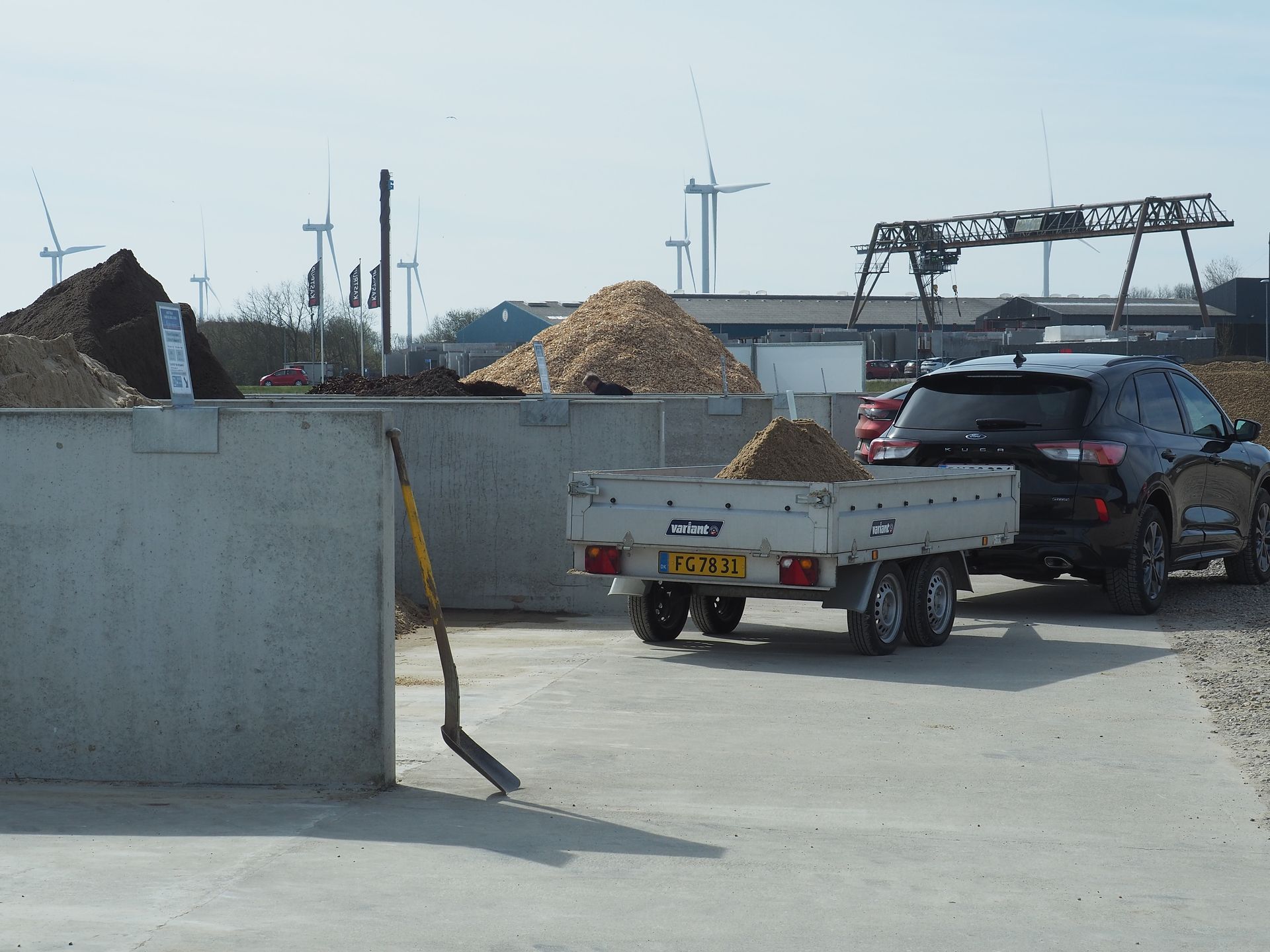 Sort SUV trækker en lille trailer læsset med grus på et udendørs anlæg; betonvægge, bunker af materiale, vindmøller.