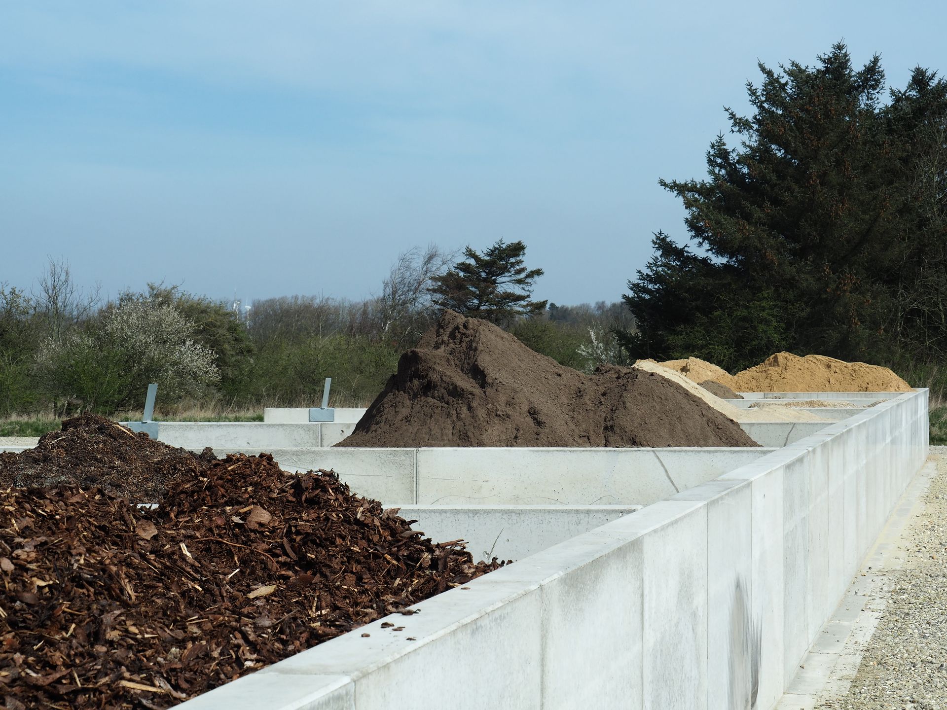 Kompostbeholdere med bunker af mørk jord, træflis og sand under en blå himmel.