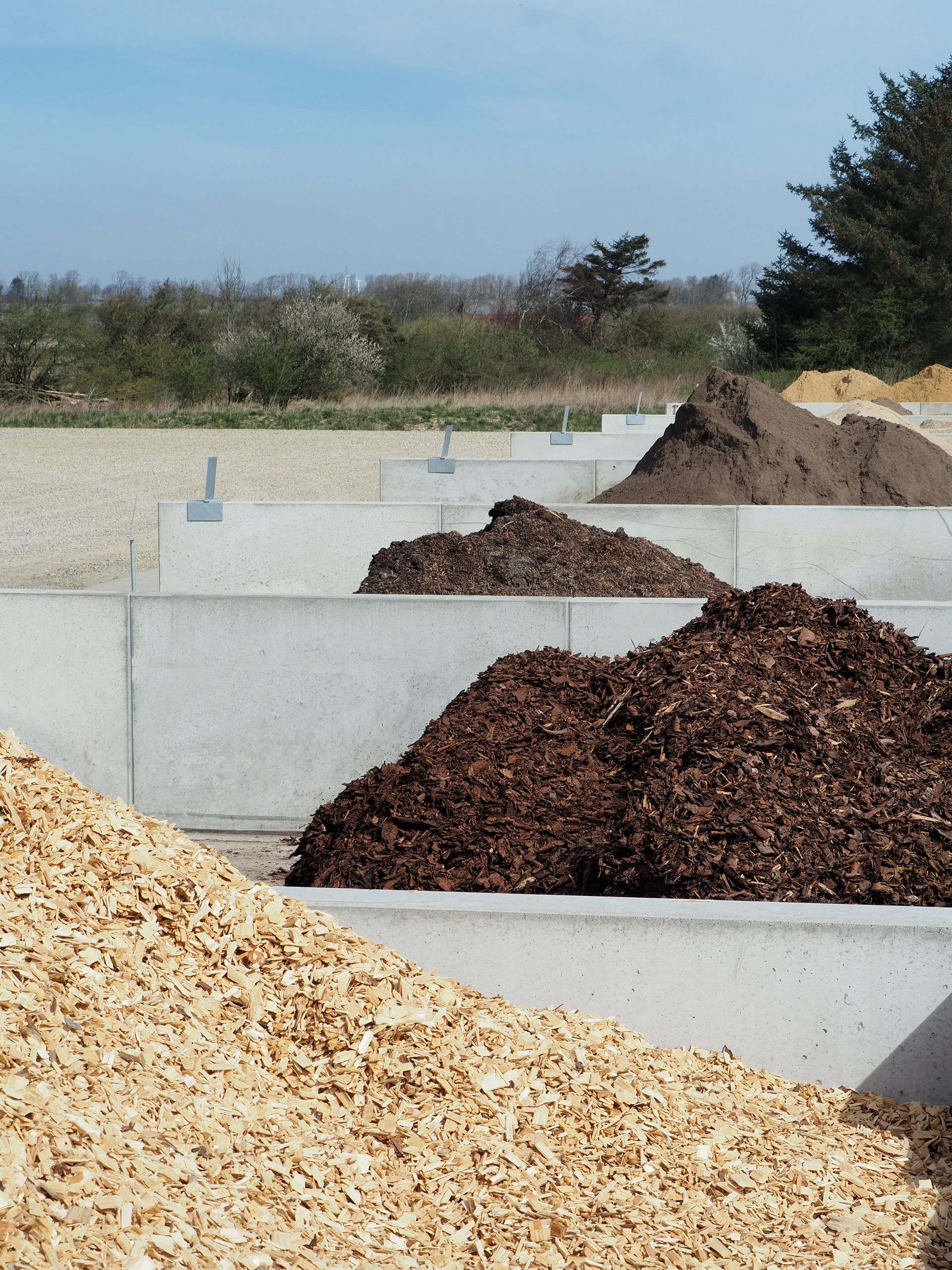 Bunker af træflis, barkflis og jord i betonbeholdere på et udendørs opbevaringsanlæg.