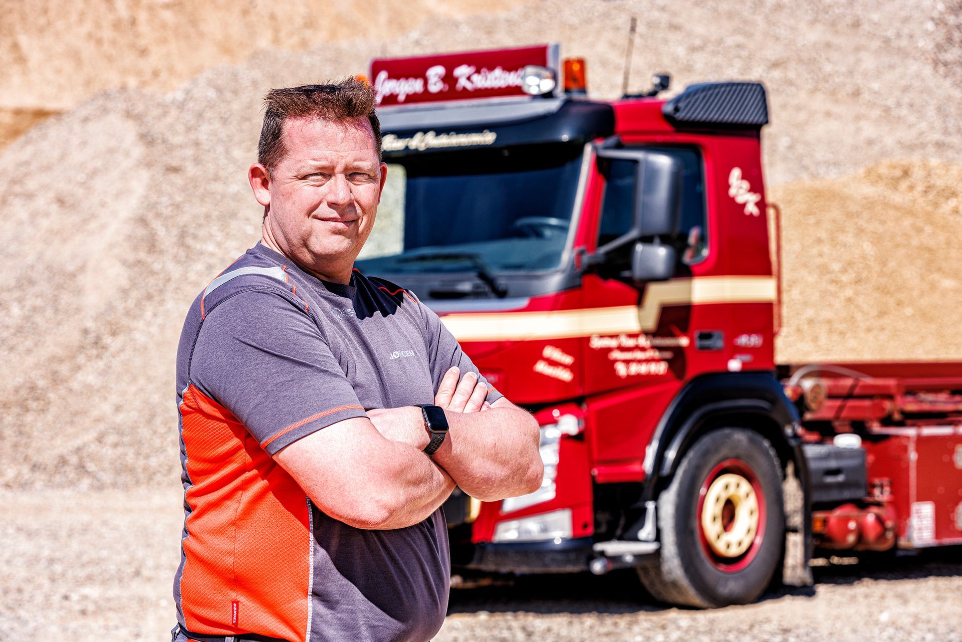 To røde og blå semitrailere parkeret udendørs på grus under en blå himmel.