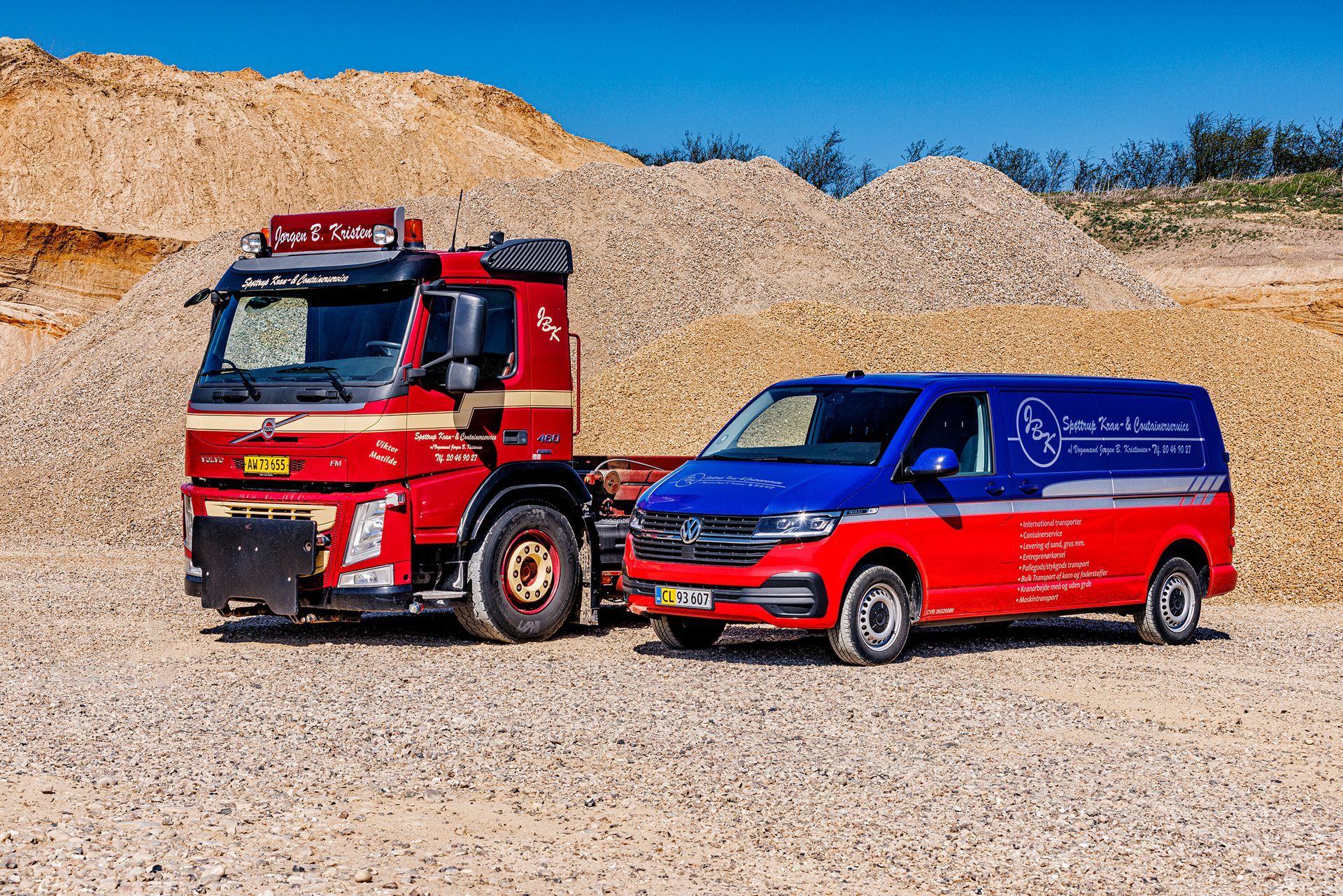 Rød Volvo lastbil og blå/rød varevogn parkeret på en grusplads med grusbunker i baggrunden.