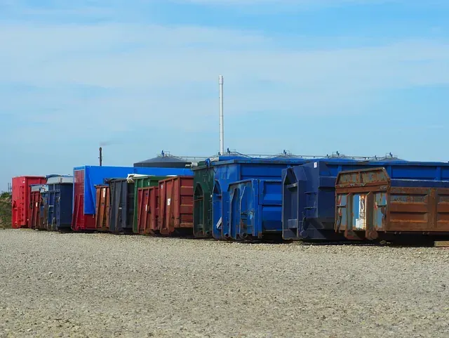 Række af farverige skraldespande på en grusplads under en blå himmel, med et tårn og en bygning i det fjerne.
