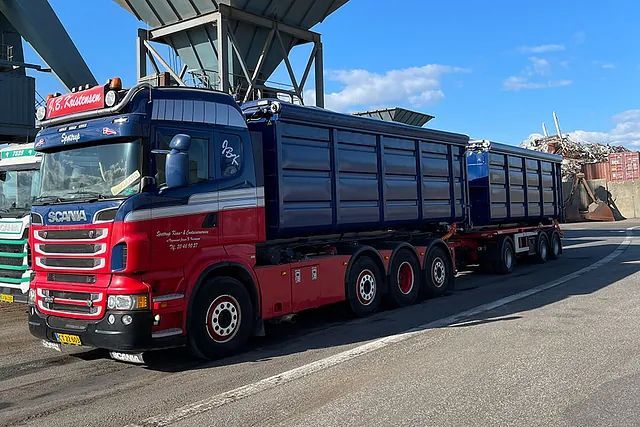 Rød og blå Scania-lastbil med trailer læsset med skraldespande i en havn under en blå himmel.