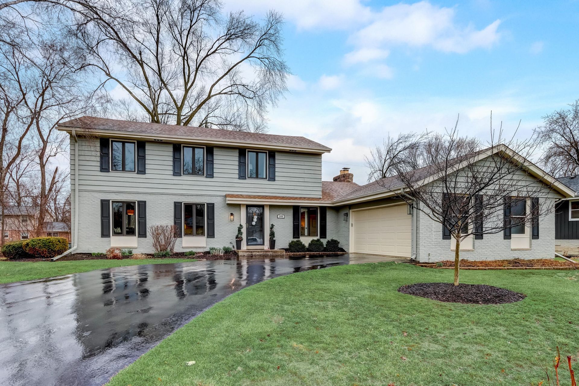 Two-story light blue house with attached garage, wet driveway, and green lawn on a cloudy day.