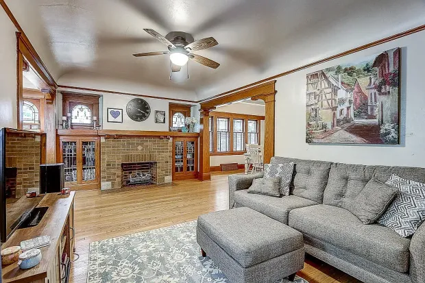 Living room with wood details, brick fireplace, gray sofa, ottoman, artwork, and ceiling fan.