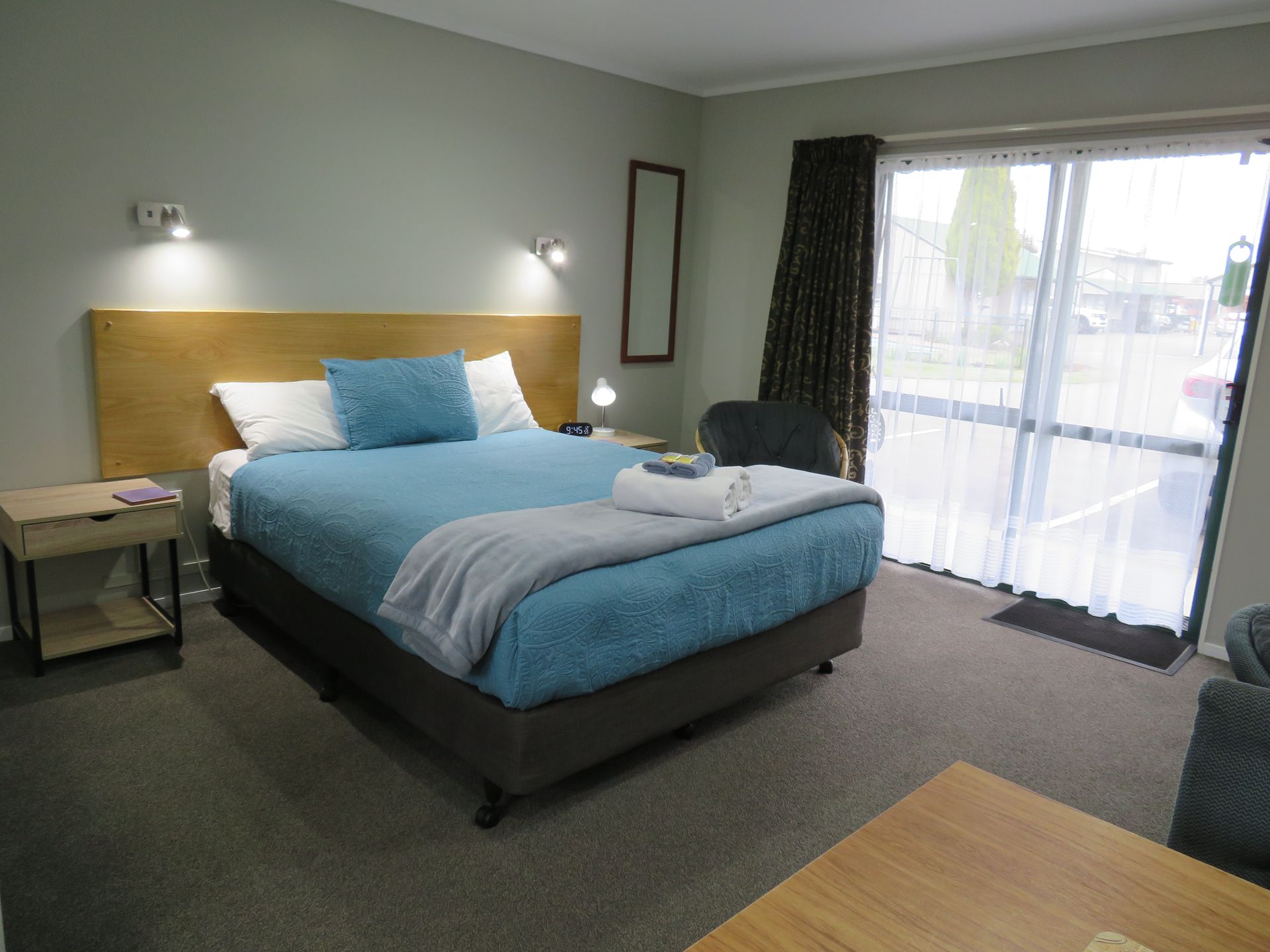 A hotel room with a bed featuring a blue comforter, light wood headboard, a bedside table, and sliding glass doors.
