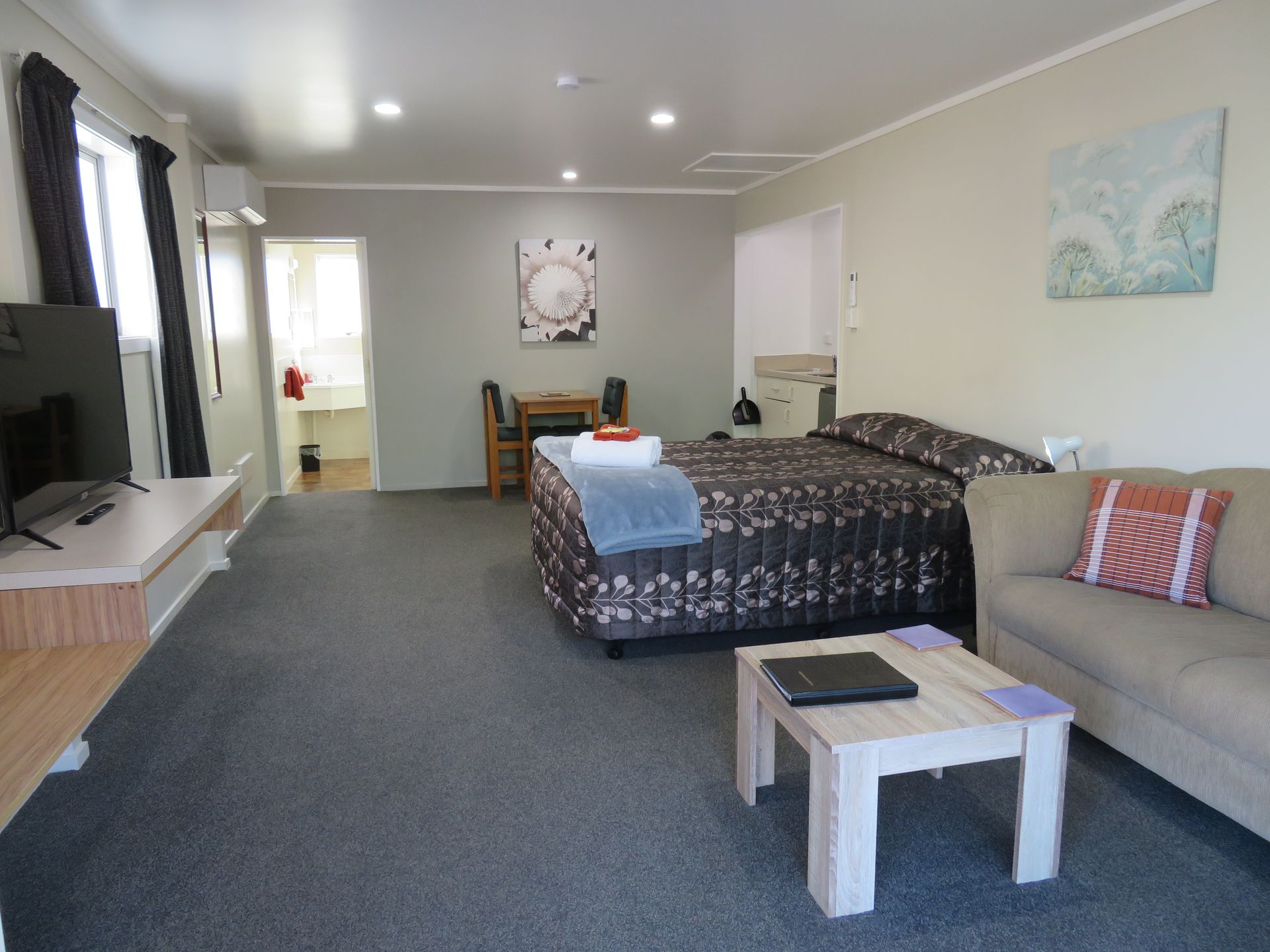 A hotel room with a queen bed, a beige couch, a square wooden coffee table, a small dining set, and a flat-screen TV.