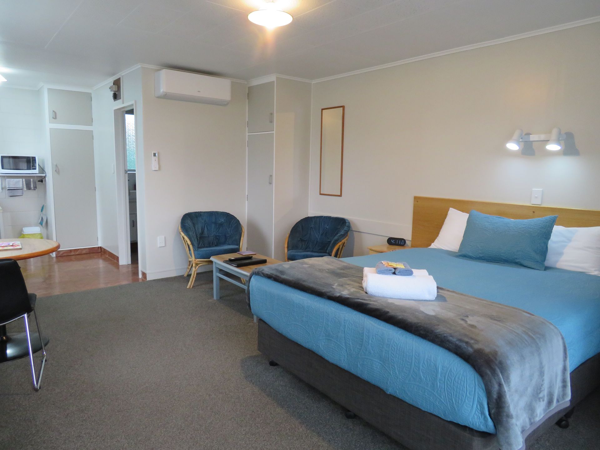 A hotel room featuring a bed with blue bedding, two armchairs, a small table, and a kitchenette area.