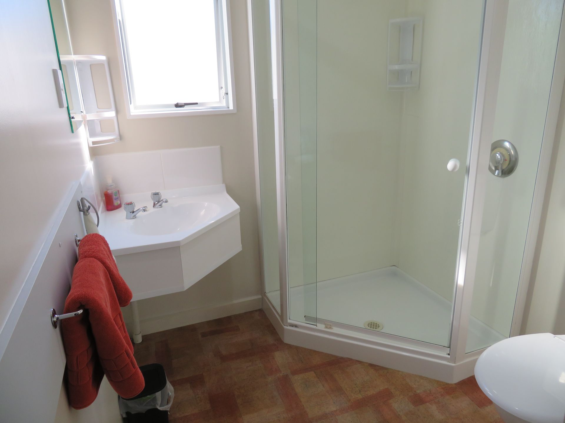A small bathroom featuring a corner sink, a glass-enclosed shower, a red towel on a rack, and a brown tiled floor.