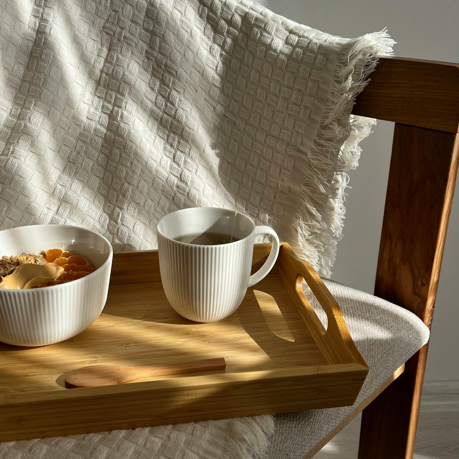 A wooden serving tray holds a white ribbed bowl of oatmeal and a matching mug on a chair with a white textured throw.