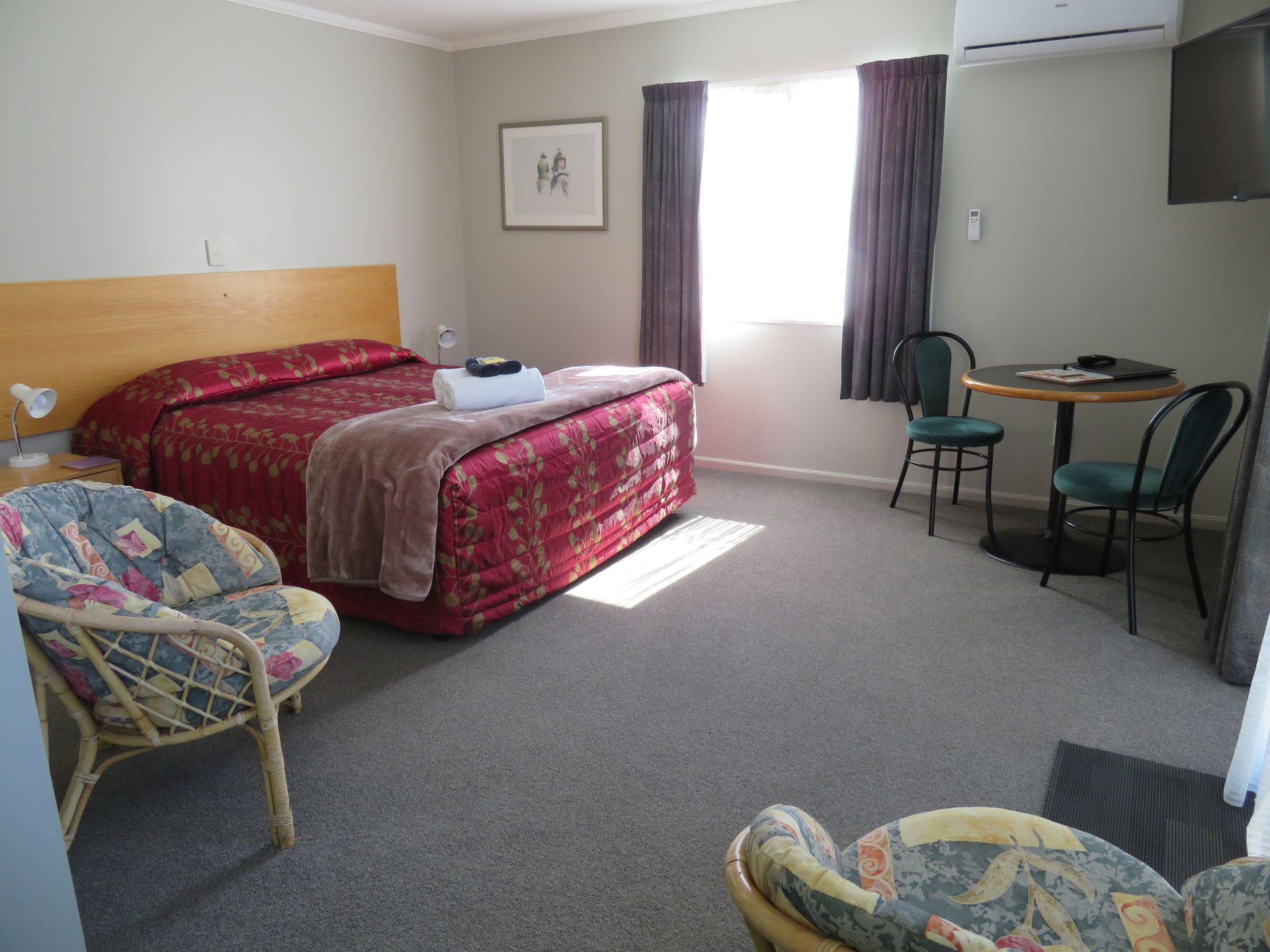 A hotel room with a queen bed featuring a red patterned bedspread, two floral armchairs, and a small dining table.
