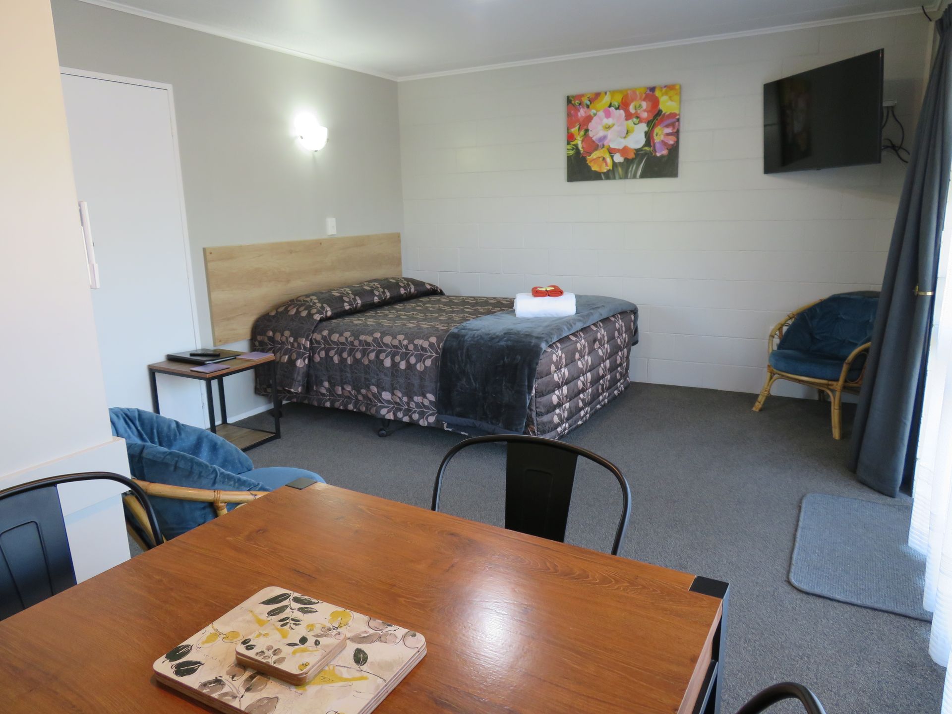 A hotel room with a queen bed, a wood dining table, two blue chairs, a wall-mounted TV, and a floral painting.
