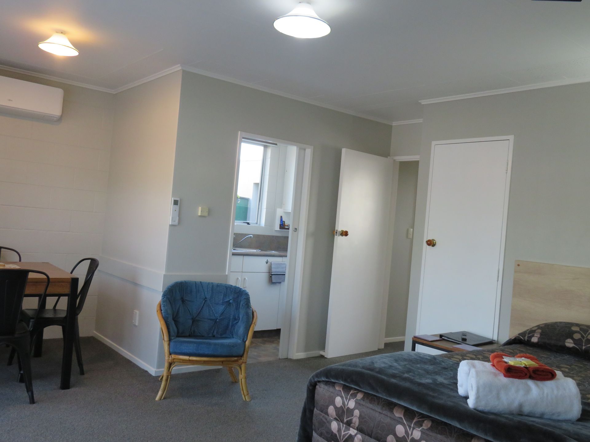 A hotel room featuring a bed with folded white towels, a blue armchair, a small dining table, and a doorway to a kitchen.