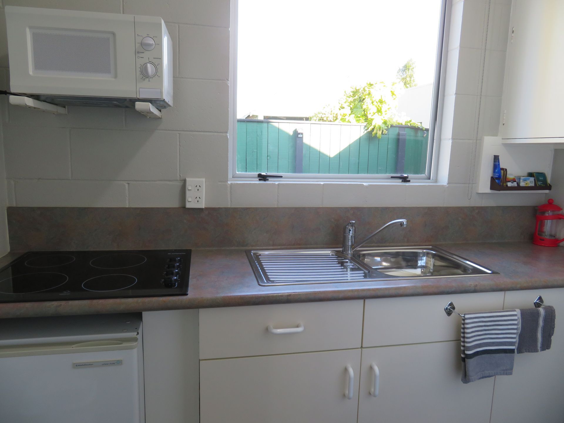 A kitchen featuring a black stovetop, stainless steel sink, white cabinets, and a microwave on a shelf beneath a window.