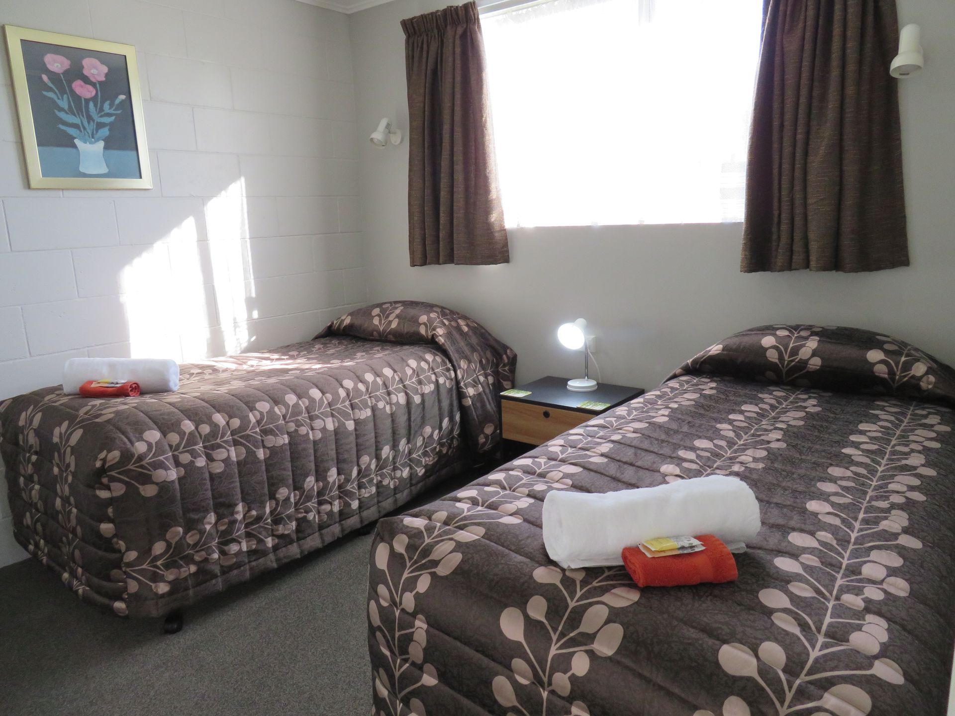 A hotel room with two twin beds featuring brown patterned bedding, separated by a nightstand with a lamp.