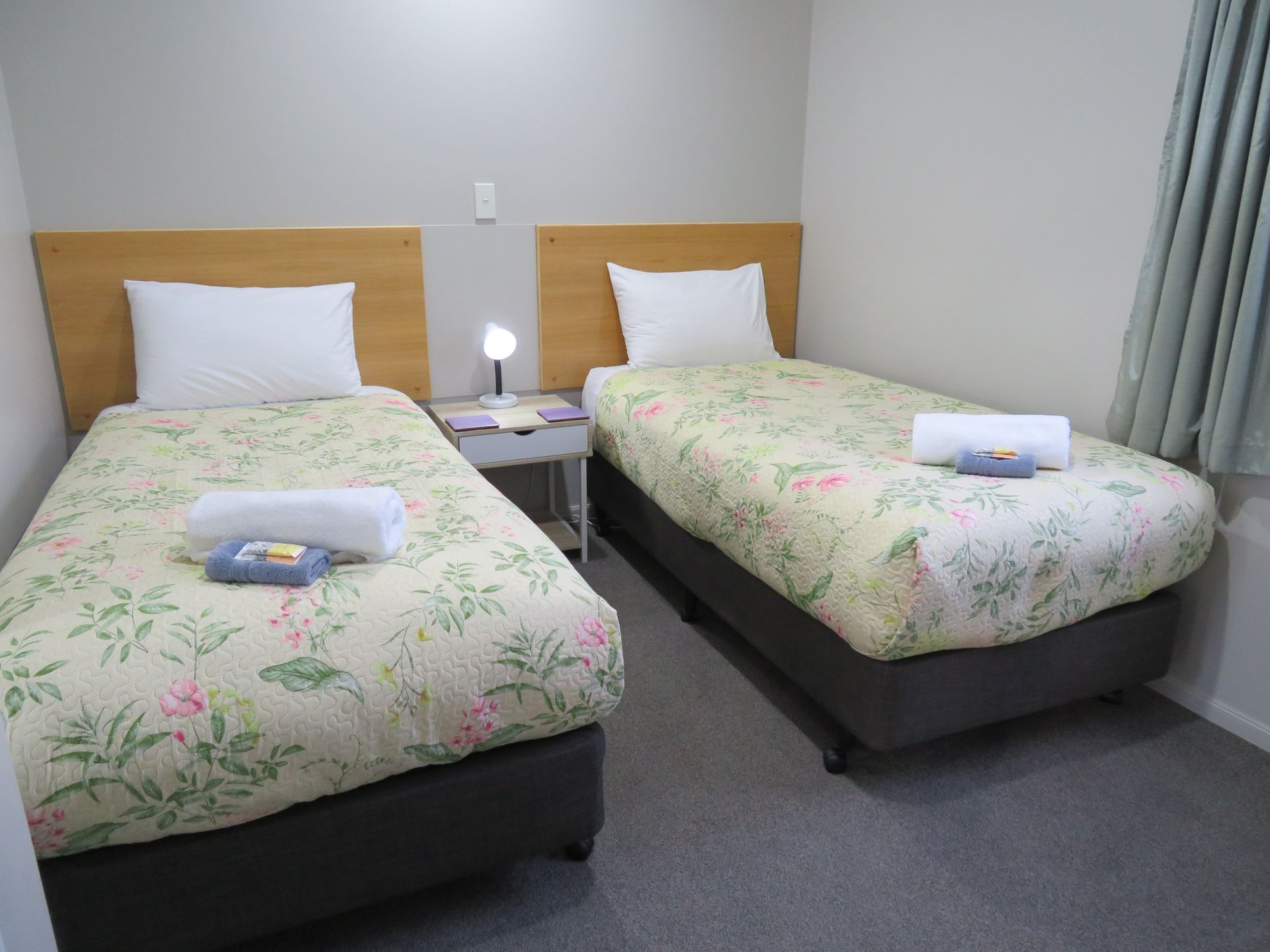 A hotel room featuring two twin beds with floral bedspreads, white pillows, folded towels, and a central bedside lamp.