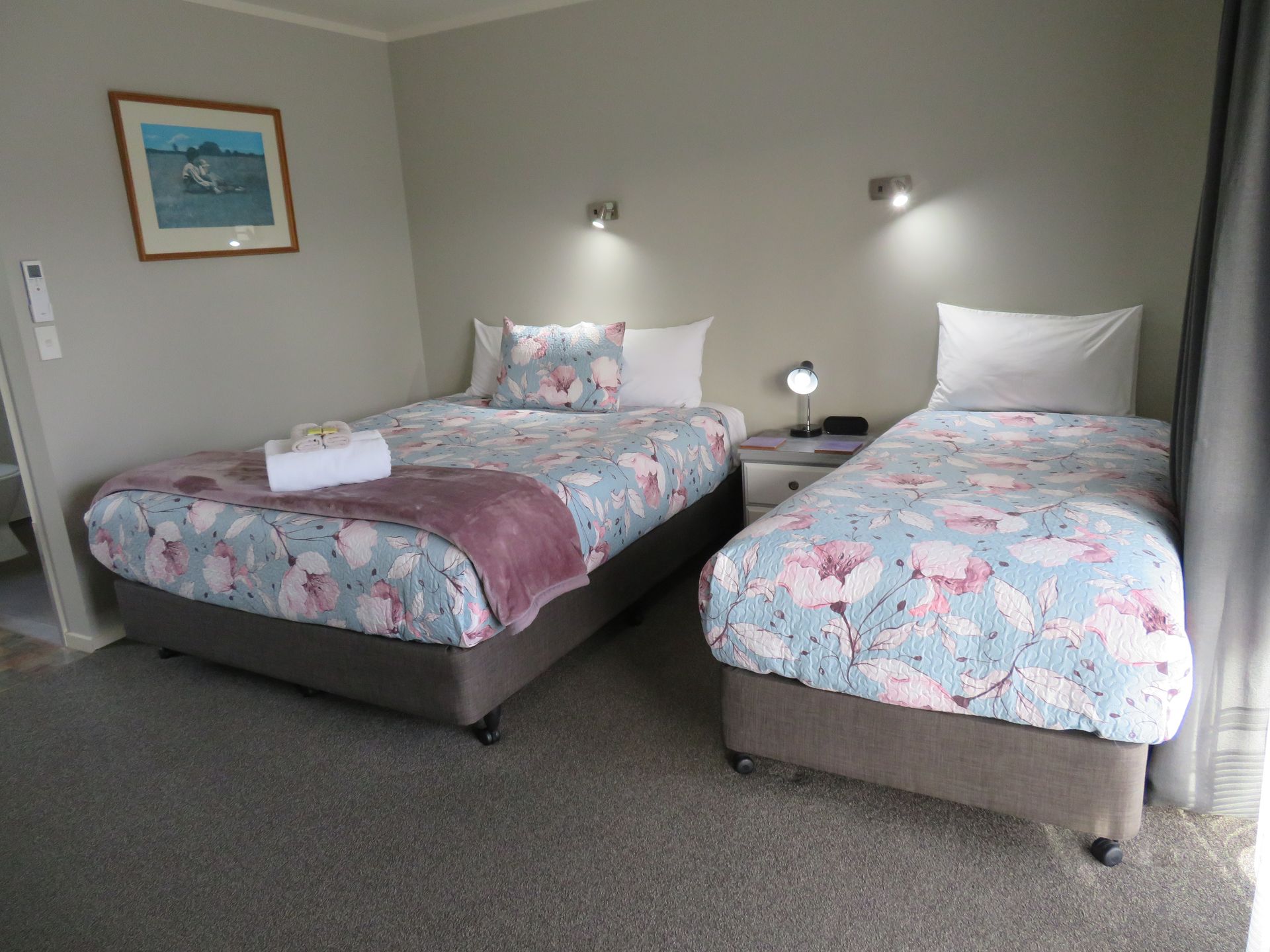 A hotel room with two beds featuring floral patterned duvets, grey carpets, neutral walls, and a small framed painting.