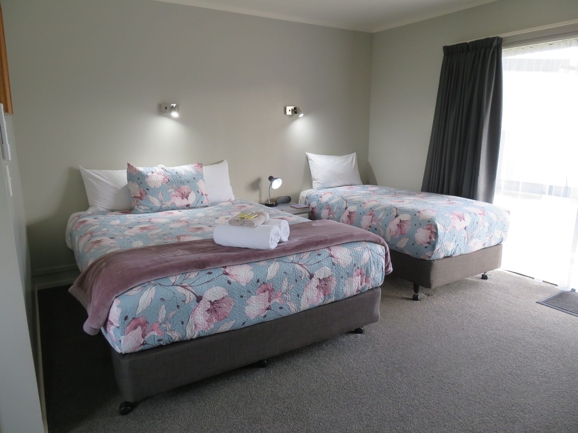 A motel room with one queen bed and one single bed, both with floral bedding, separated by a bedside table and lamp.