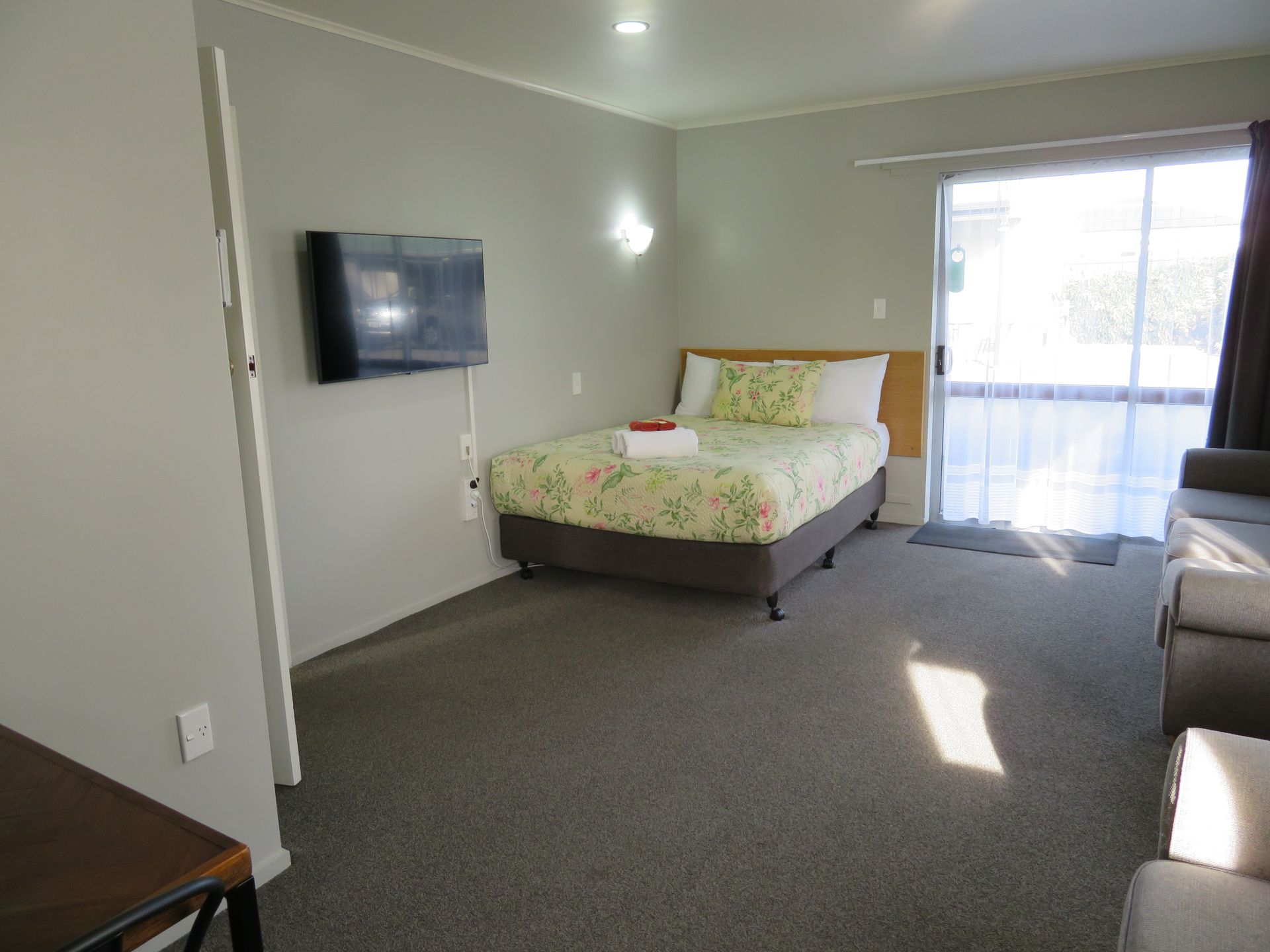 A furnished hotel room with a double bed, a wall-mounted TV, a light gray carpet, and a large sliding glass door.