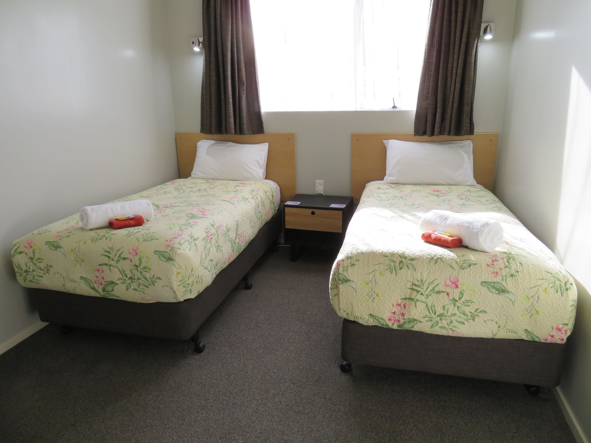 A hotel room with two twin beds featuring floral bedding, white pillows, brown curtains, and a central nightstand.