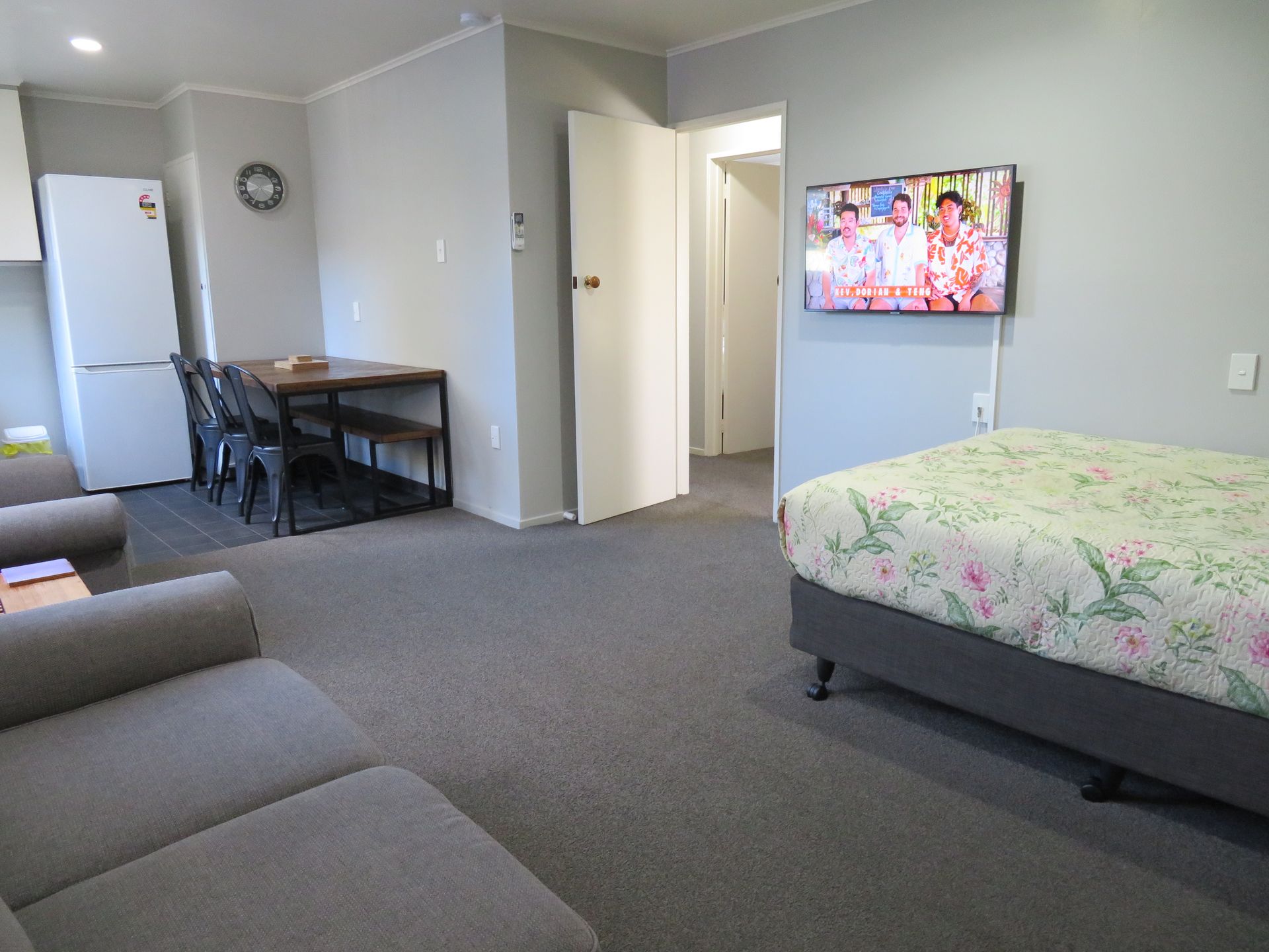 A studio apartment featuring a bed with a floral comforter, a sofa, a dining table with chairs, and a wall-mounted TV.