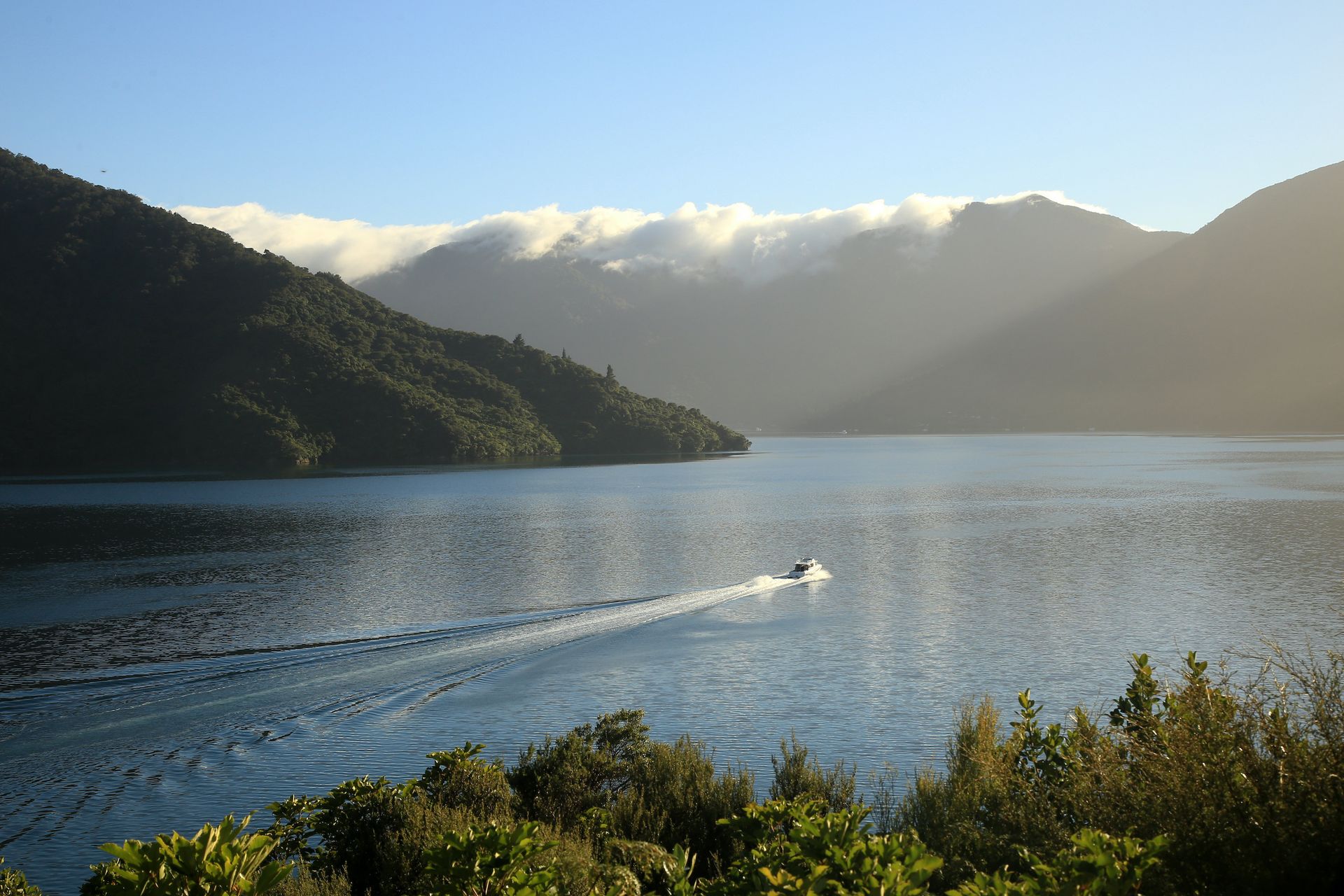 A boat leaves a wake on a calm lake surrounded by mountains under a bright, sunny sky with low-hanging clouds.