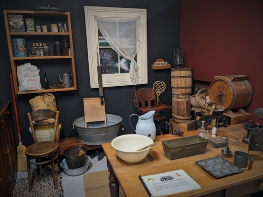 A historical kitchen display featuring wooden shelves, a wash basin, a butter churn, and antique items on a table.